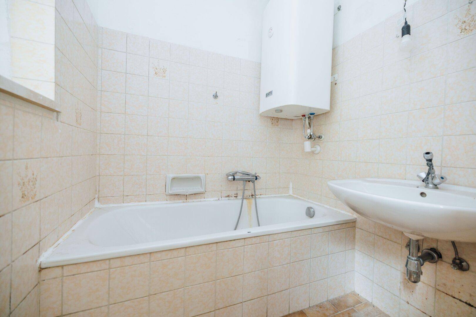 Badezimmer mit Badewanne, Waschbecken und Wandfliesen in hellem Beige, mit Warmwasserboiler.