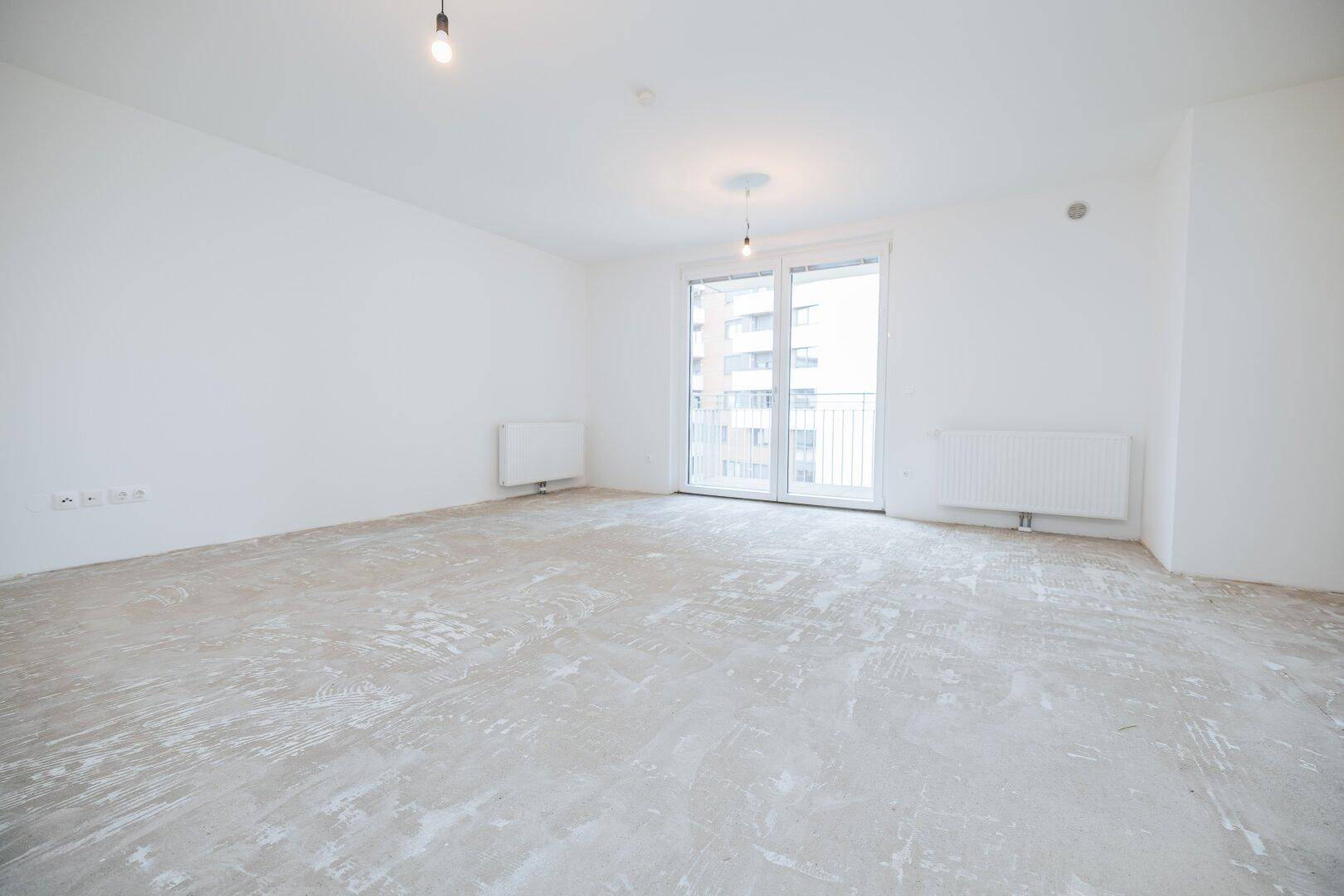 Lichtdurchfluteter Raum mit weiß gestrichenen Wänden, Betonboden und Fenster mit französischem Balkon.