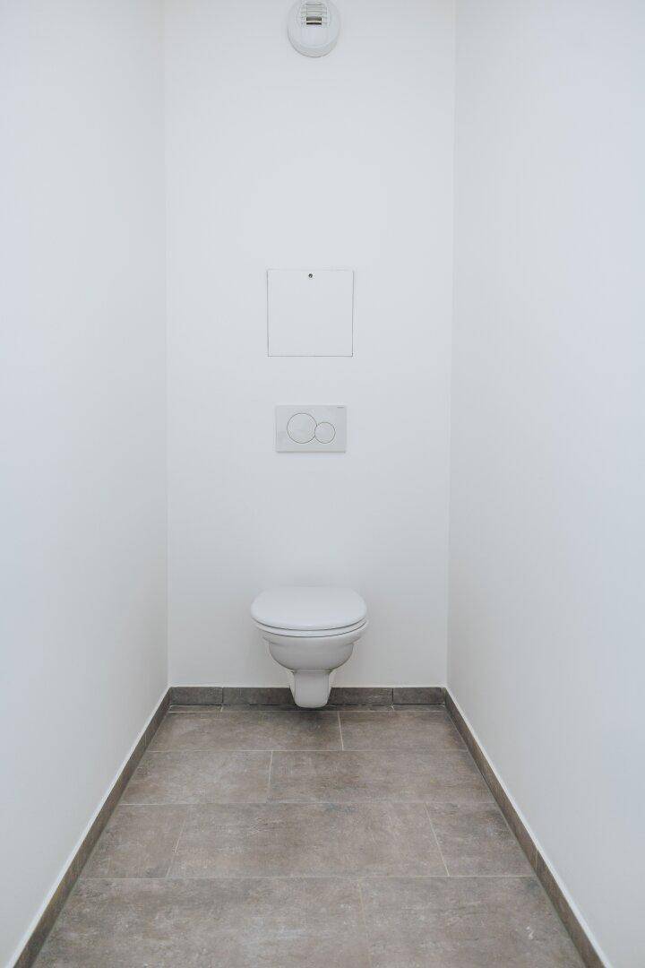 Separates WC mit weißen Wänden, grauen Bodenfliesen und wandhängender Toilette.
