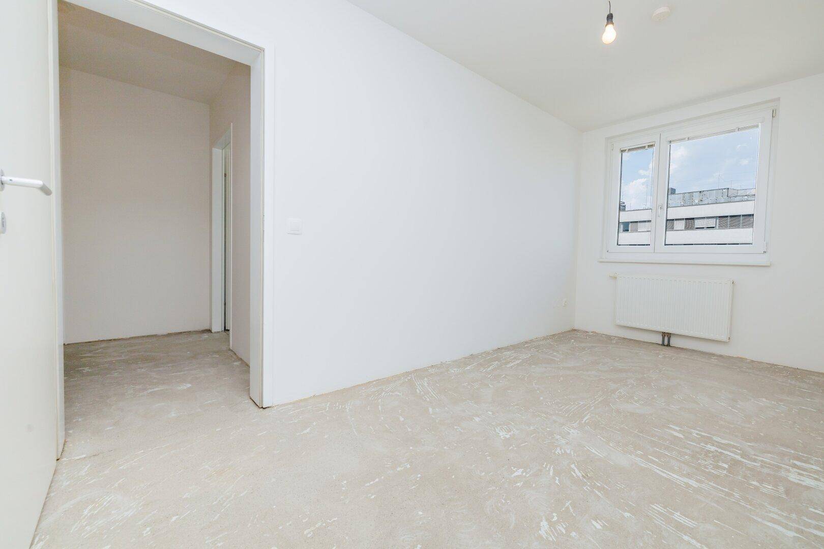 Helles Schlafzimmer mit weiß gestrichenen Wänden, Betonboden und einem Fenster.