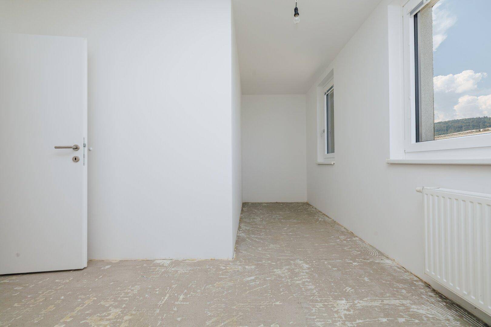 Unmöblierter Raum mit weiß gestrichenen Wänden, Betonboden und zwei Fenstern.