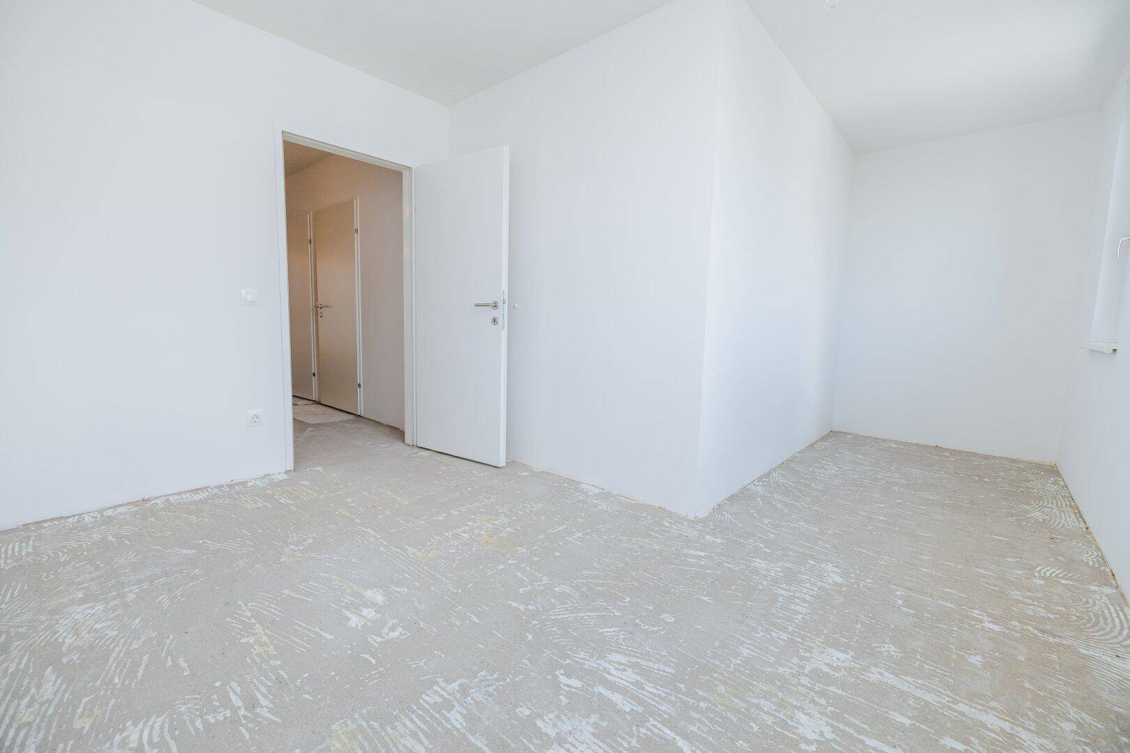 Heller Raum mit weiß gestrichenen Wänden, Betonboden und offener Tür zu einem weiteren Zimmer.