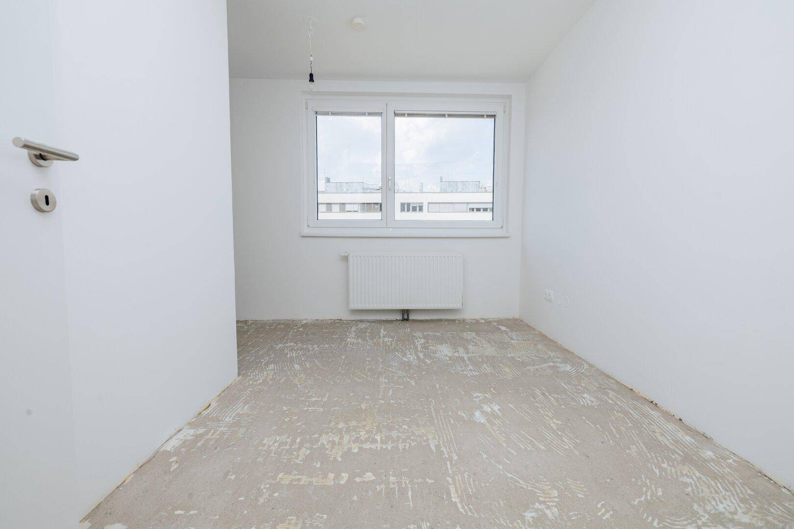 Unmöbliertes Schlafzimmer mit weiß gestrichenen Wänden, Betonboden und einem Fenster.