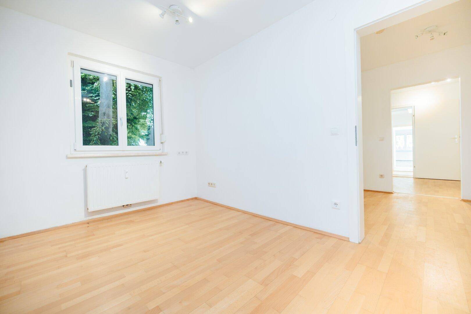 Heller Raum mit Holzboden und Fenster, der einen offenen Übergang zu einem weiteren Bereich bietet.