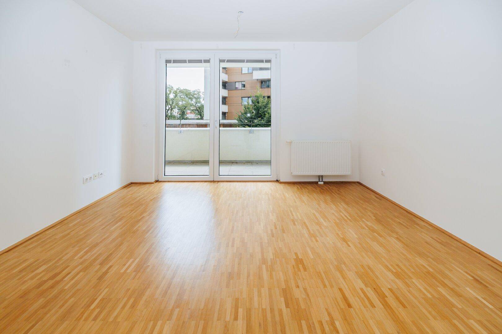 Geräumiges Zimmer mit hellem Parkettboden und großen Fenstern zum Balkon.