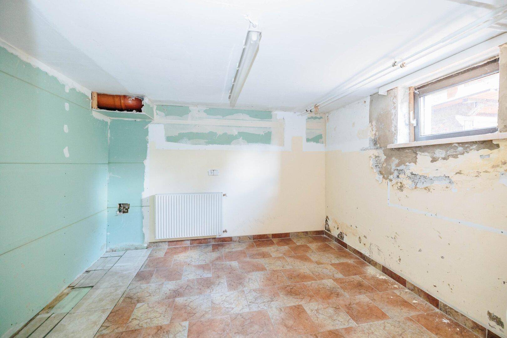 Renovierungsbedürftiger Kellerraum mit Fenster, Heizkörper und Fliesenboden.