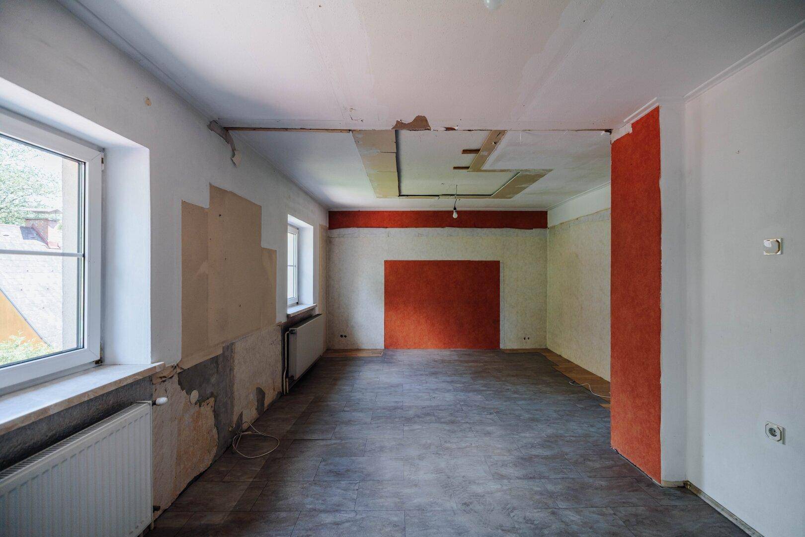 Renovierungsbedürftiges Wohnzimmer mit Fliesenboden, farbiger Akzentwand und Blick ins Grüne.