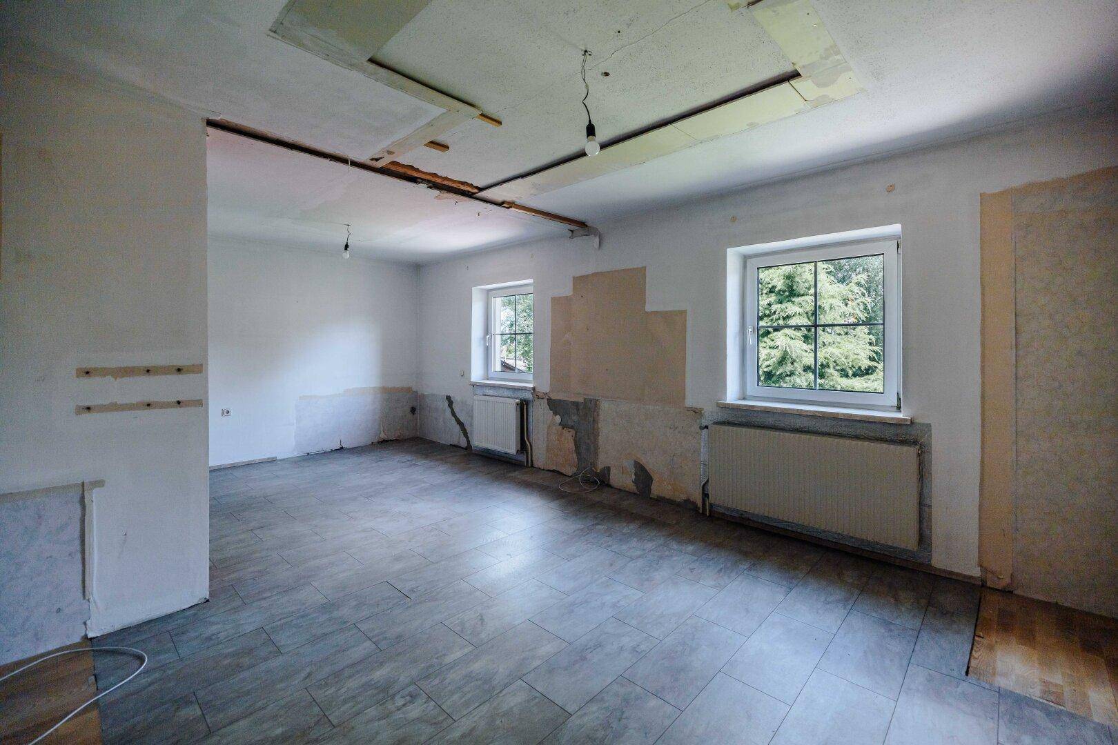 Geräumiges Wohnzimmer in Renovierung mit Fliesenboden und zwei Fenstern zum Garten.