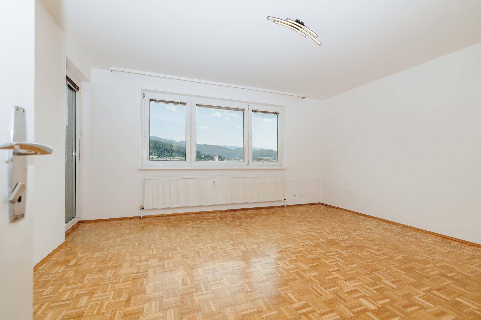 Geräumiges Zimmer mit hellem Parkettboden und weitem Blick auf die grüne Umgebung und Berge.