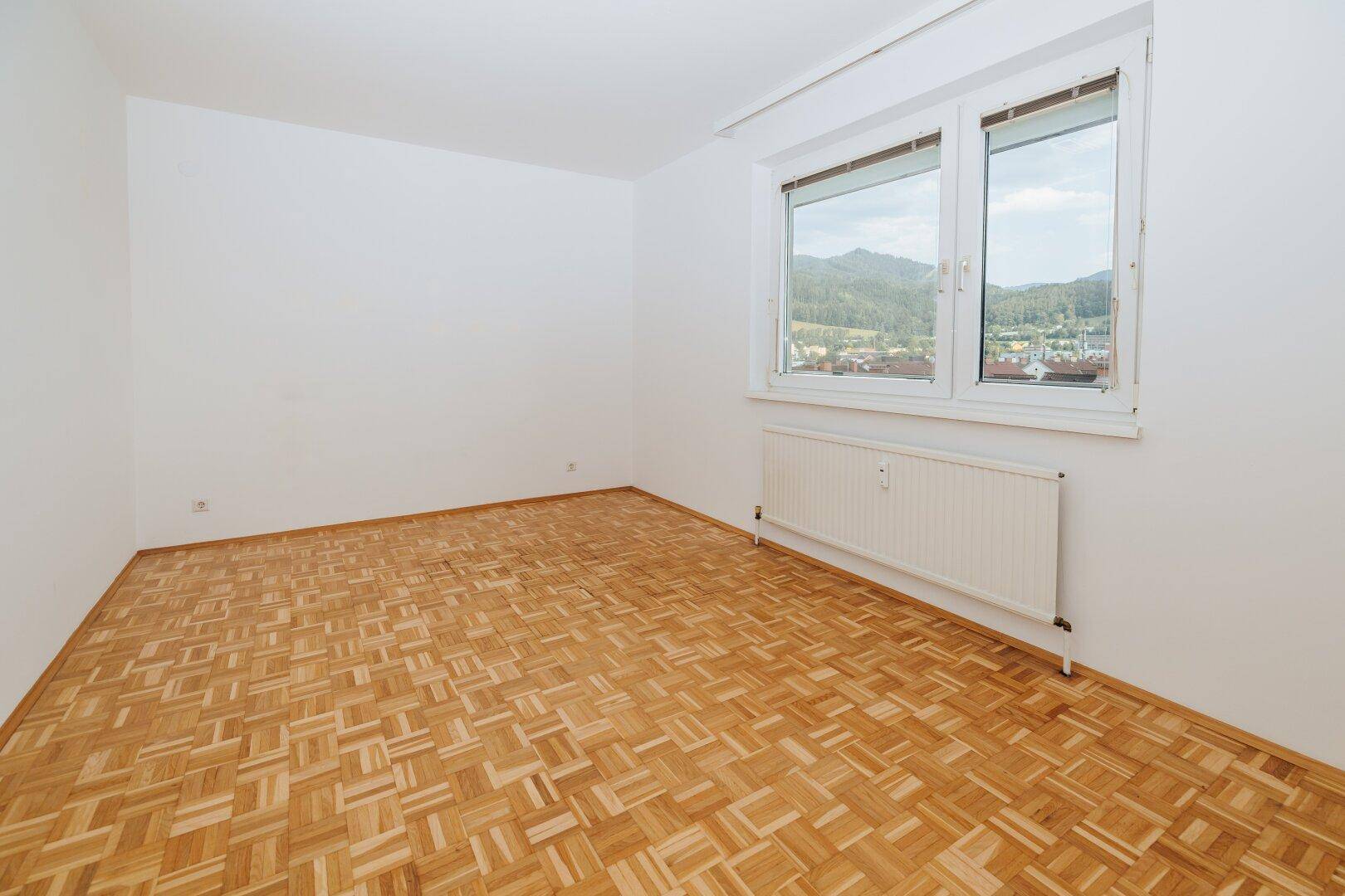 Helles Zimmer mit Parkettboden und zwei Fenstern, die einen angenehmen Blick ins Grüne ermöglichen.