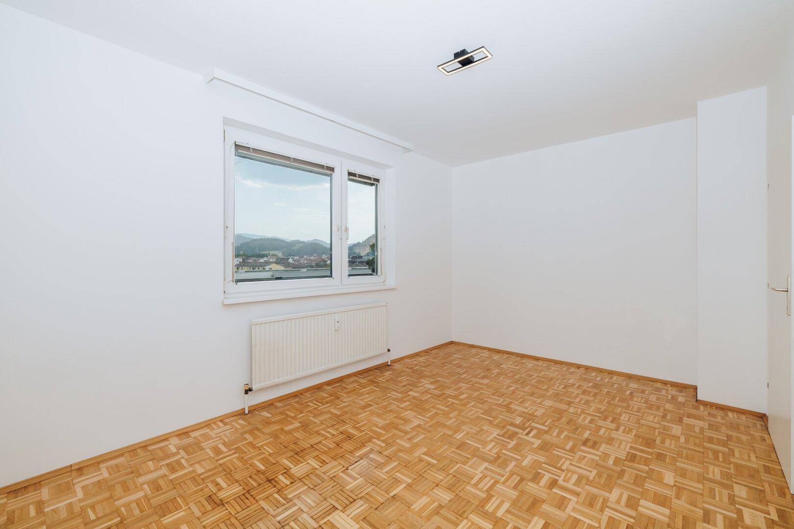 Freundliches Zimmer mit Parkettboden und Fensterfront, die viel Tageslicht und eine schöne Aussicht bietet.