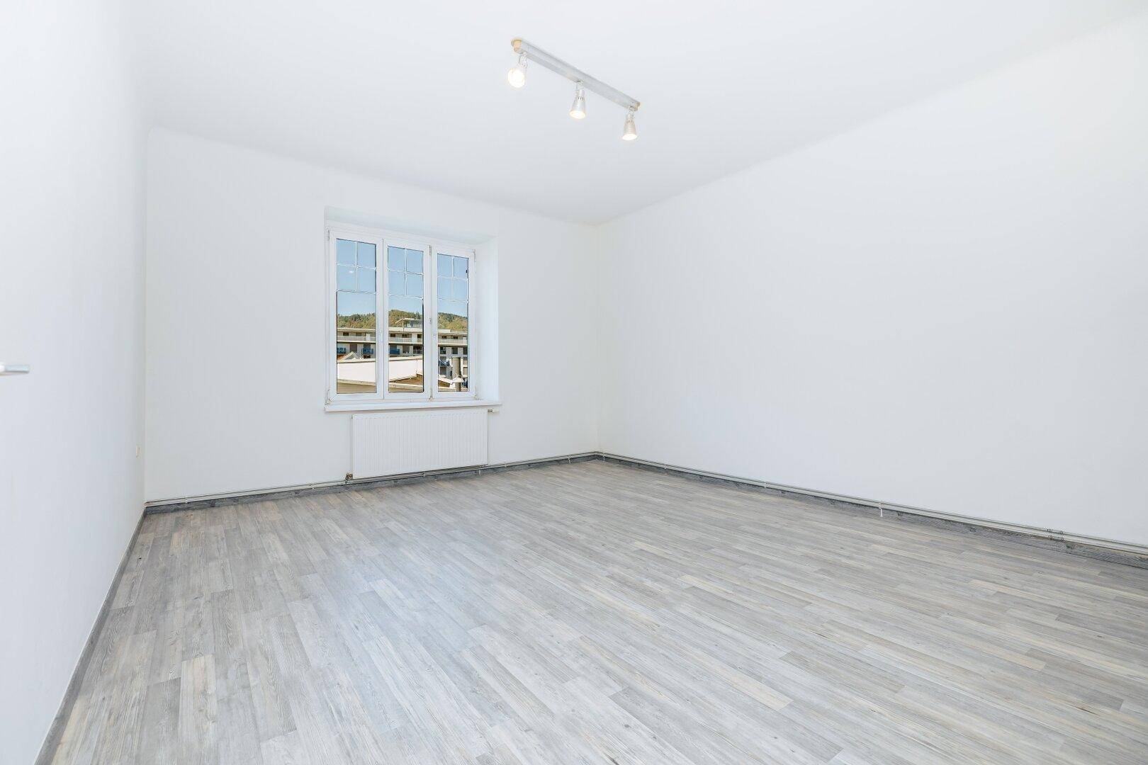 Heller, leerer Raum mit großem Fenster und modernem Laminatboden, bereit zur individuellen Gestaltung.