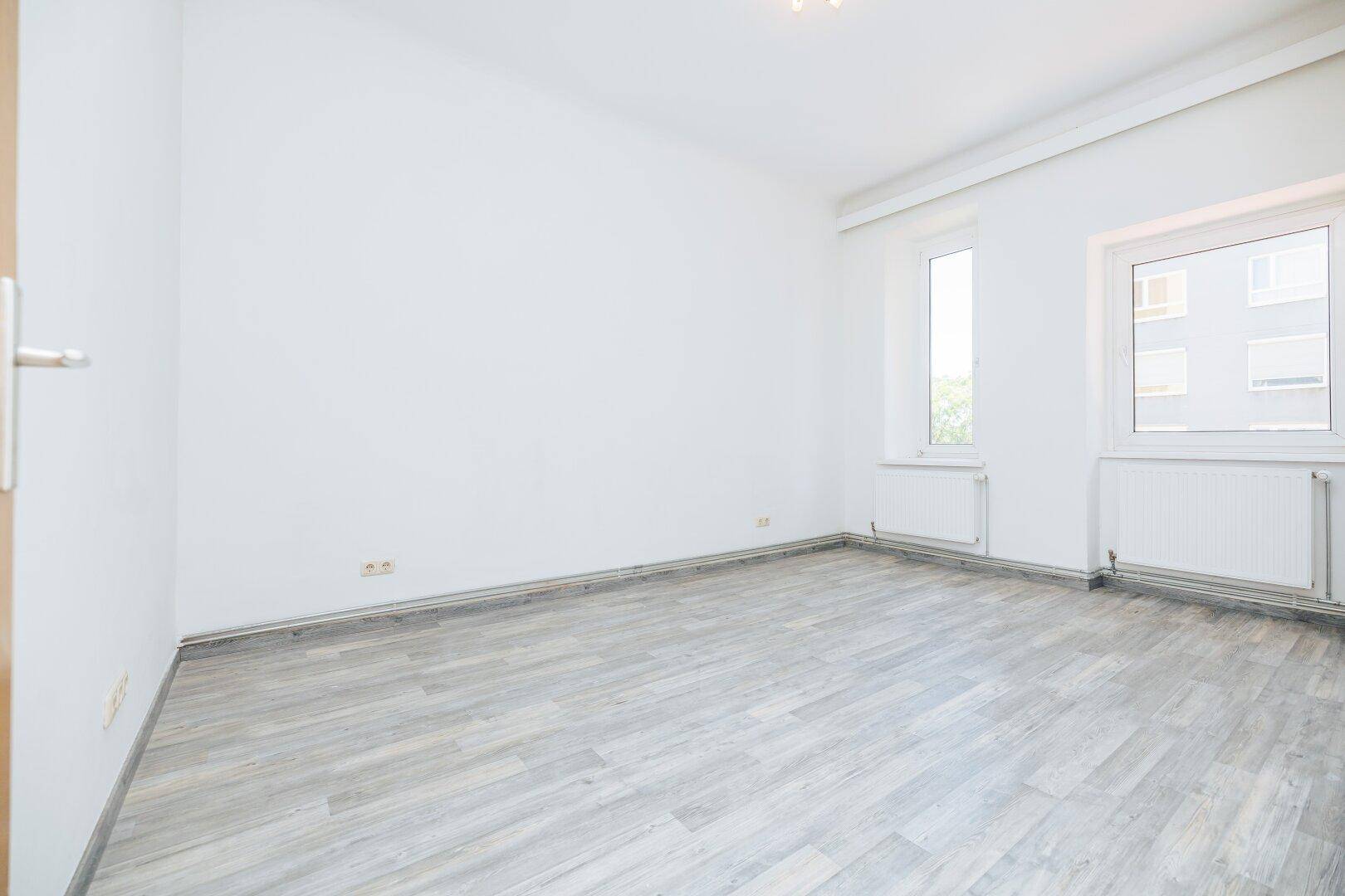Geräumiger, heller Raum mit zwei Fenstern und hellem Laminatboden, ideal für Schlaf- oder Wohnbereich.