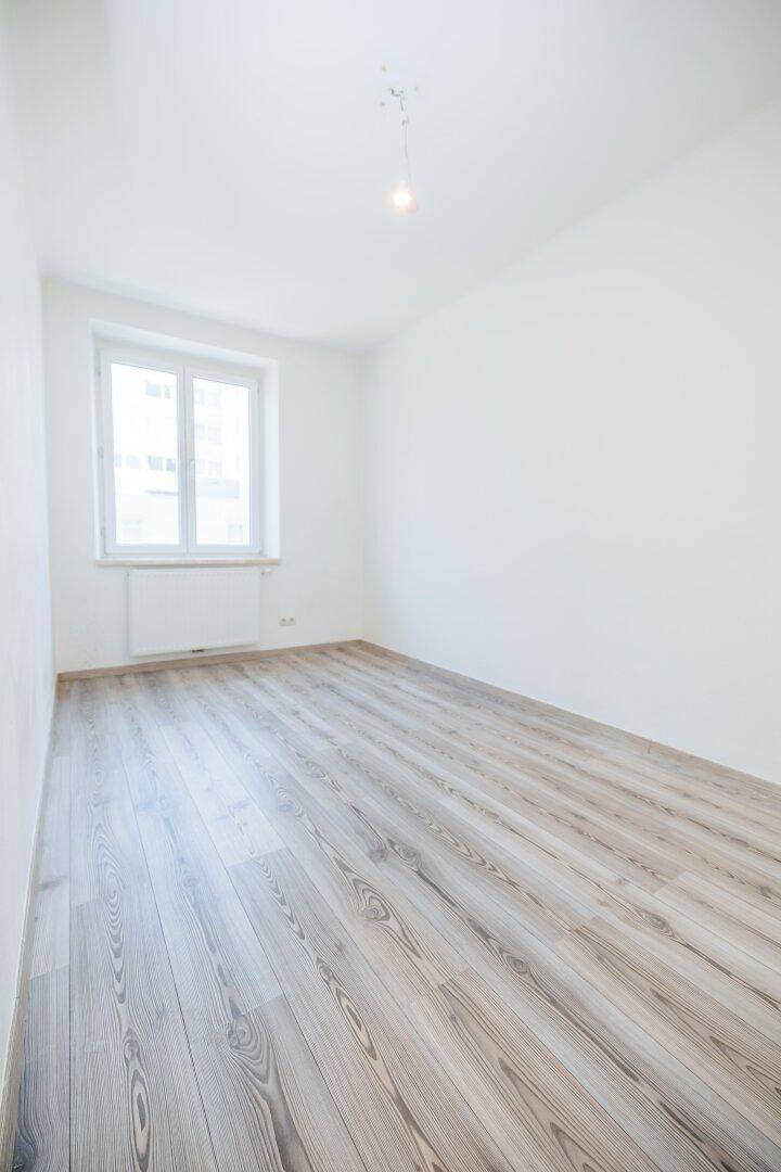 Leerer Raum mit hellem Laminatboden und einem Fenster, das Tageslicht hereinlässt.