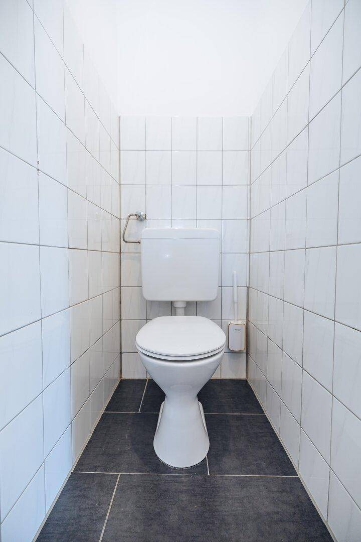 Kleines WC mit weißer Toilette und weißen Wandfliesen sowie dunklen Bodenfliesen.