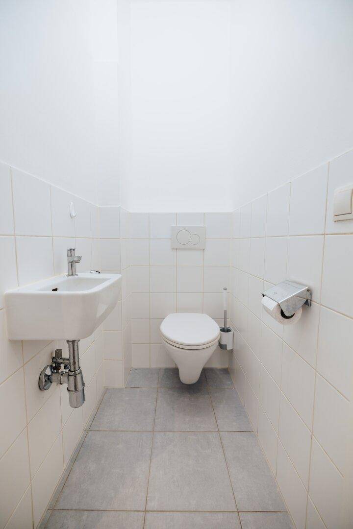 Separates WC mit wandhängender Toilette und kleinem Waschbecken, hell gefliest.