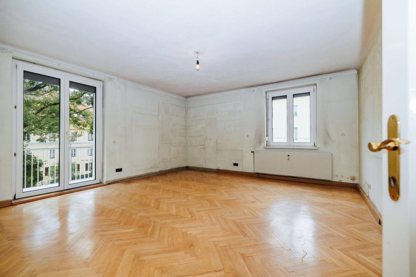 Geräumiges Wohnzimmer mit Fischgrätparkett, großem Fenster und Zugang zum Balkon.