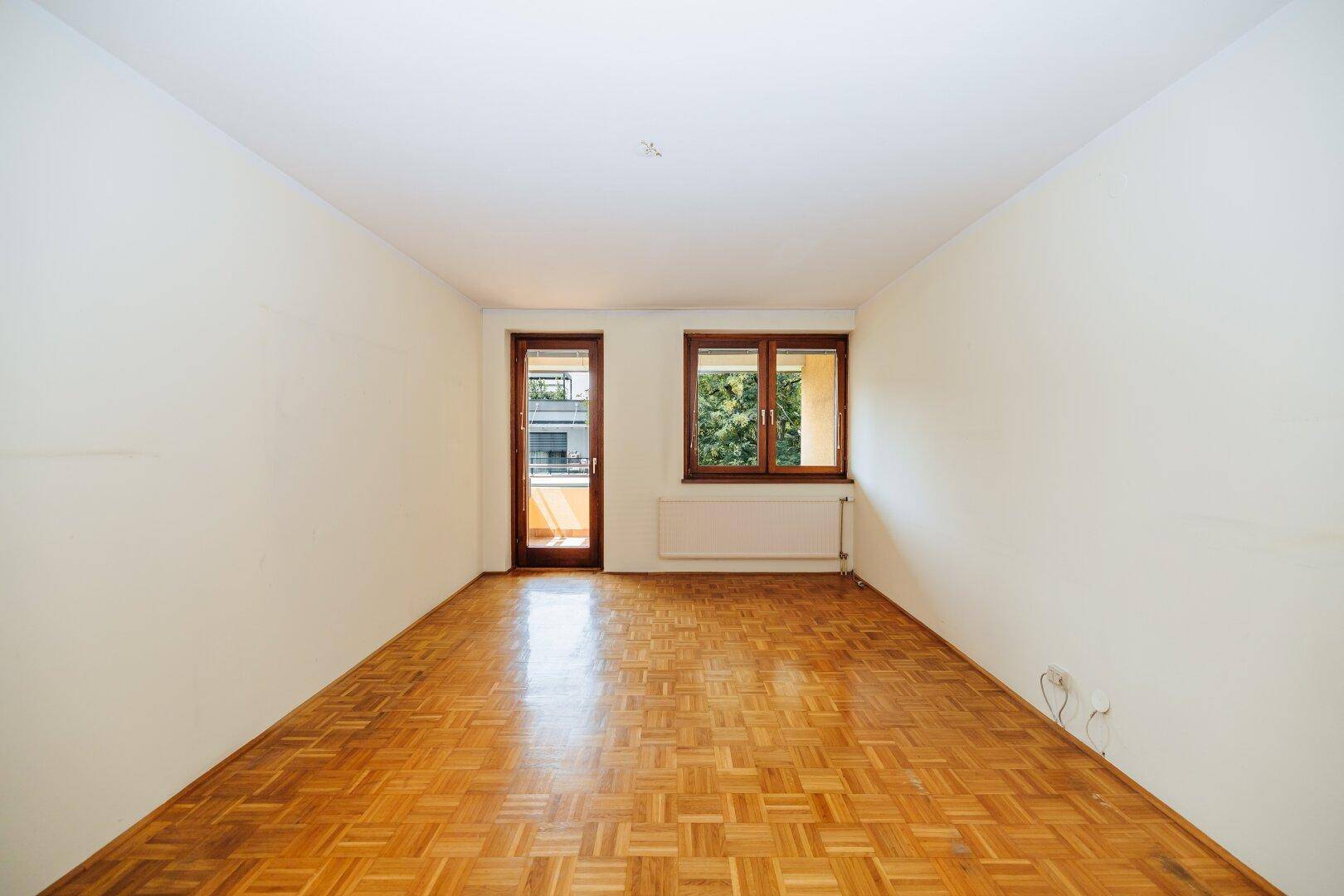 Helles, leeres Zimmer mit Fischgrätparkett, weißen Wänden und Zugang zu einem Balkon.