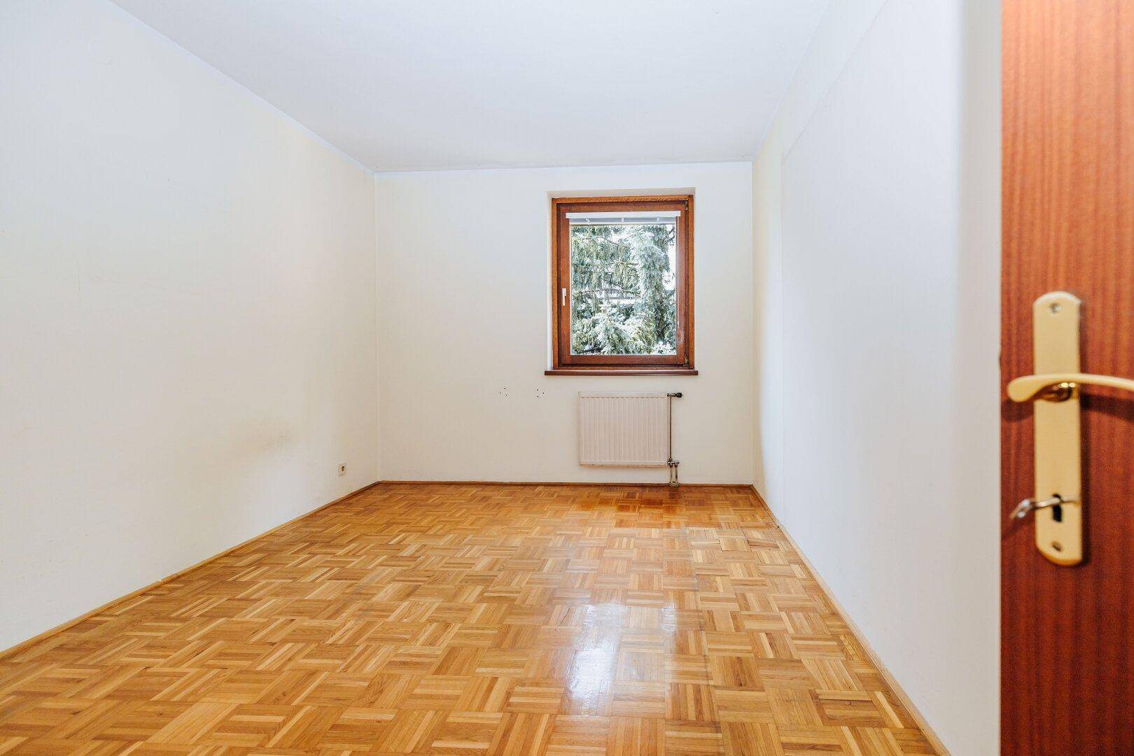 Freundliches Zimmer mit Parkettboden, weißen Wänden und einem Fenster, das viel Tageslicht hereinlässt.