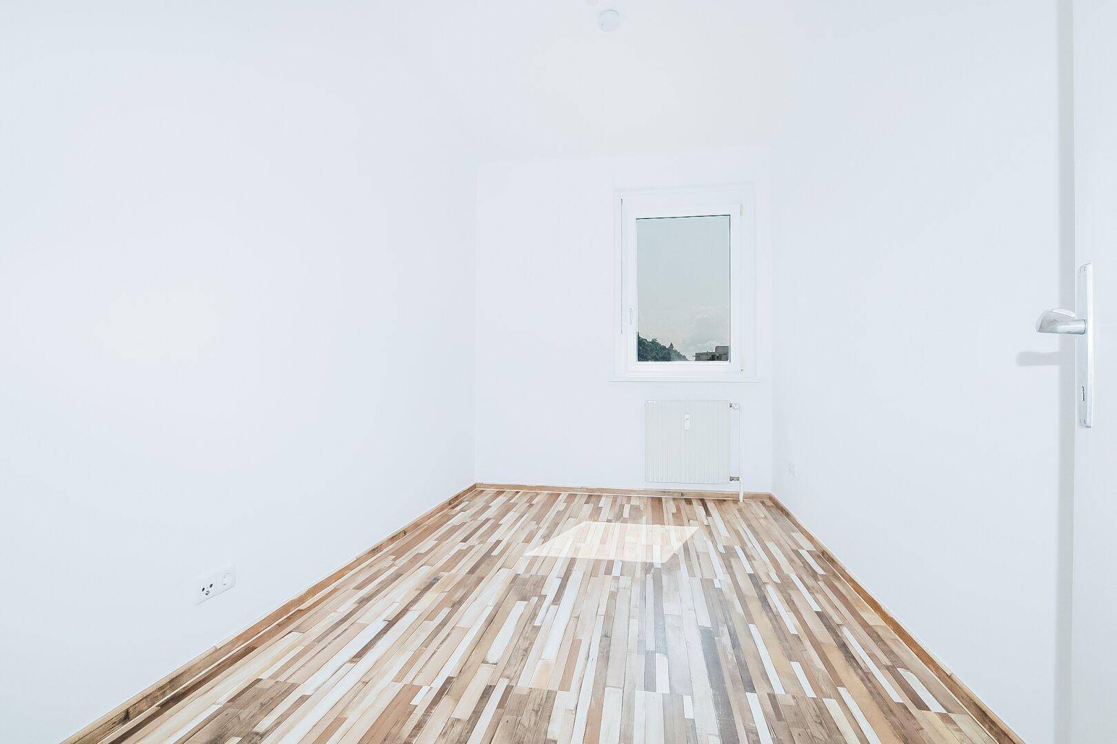 Leeres Zimmer mit hellem Laminatboden und einem Fenster, bereit zur individuellen Gestaltung.