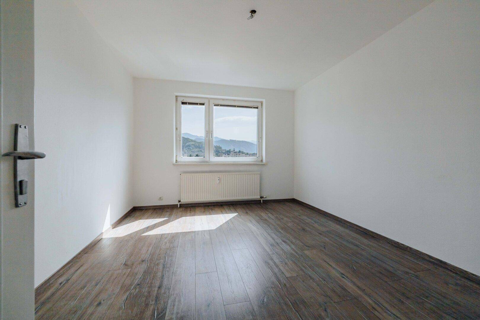 Geräumiges Zimmer mit Laminatboden, weiß gestrichenen Wänden und Ausblick in die Natur.