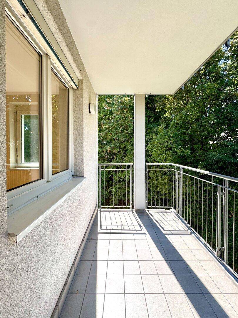 Sonniger Balkon mit Fliesenboden und Blick ins Grüne, umgeben von Bäumen.