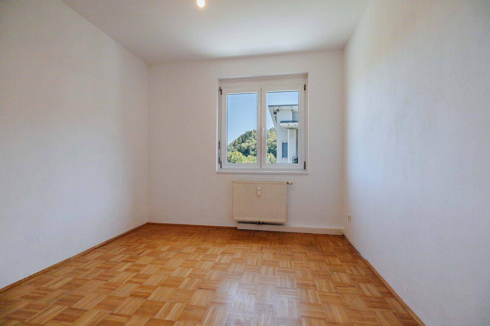 Helles Schlafzimmer mit Parkettboden, weißen Wänden und Fenster mit Ausblick.