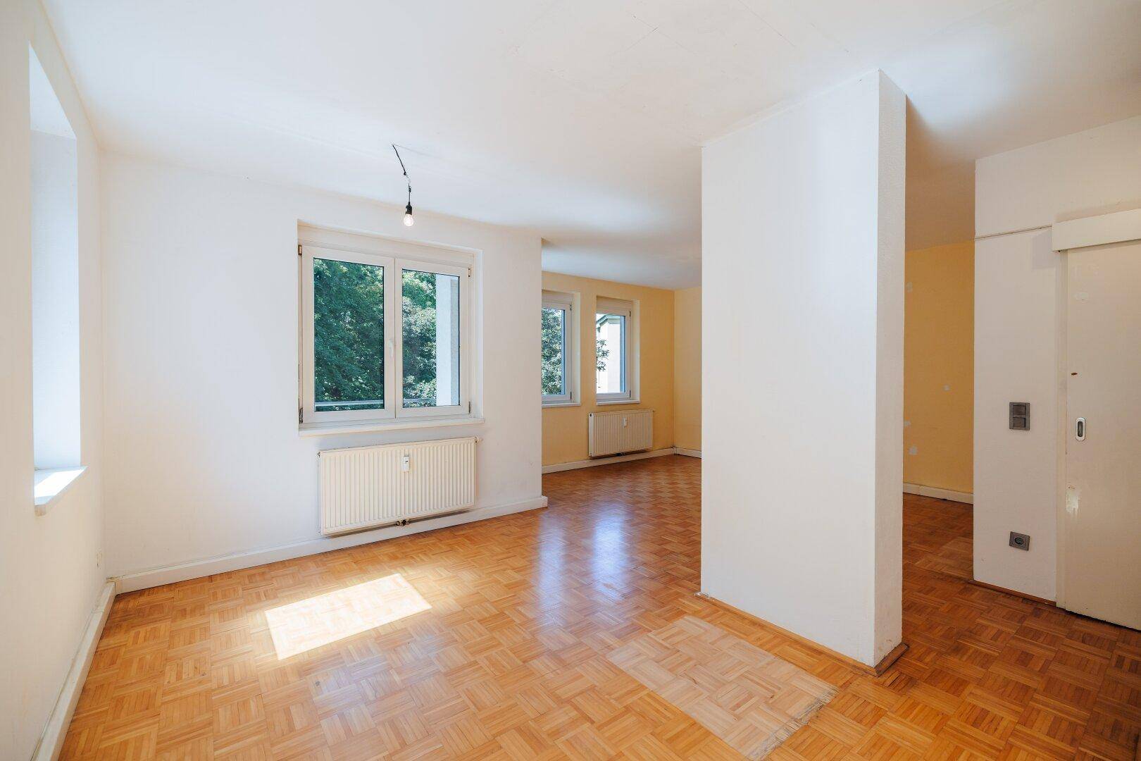 Heller Wohnraum mit Parkettboden, großen Fenstern und einem strukturellen Pfeiler.