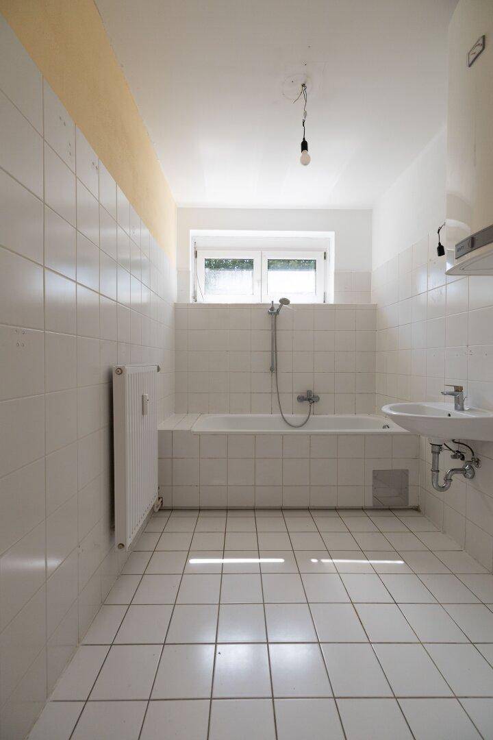 Badezimmer mit weißen Fliesen, Badewanne mit Duschfunktion und Fenster für Tageslicht.