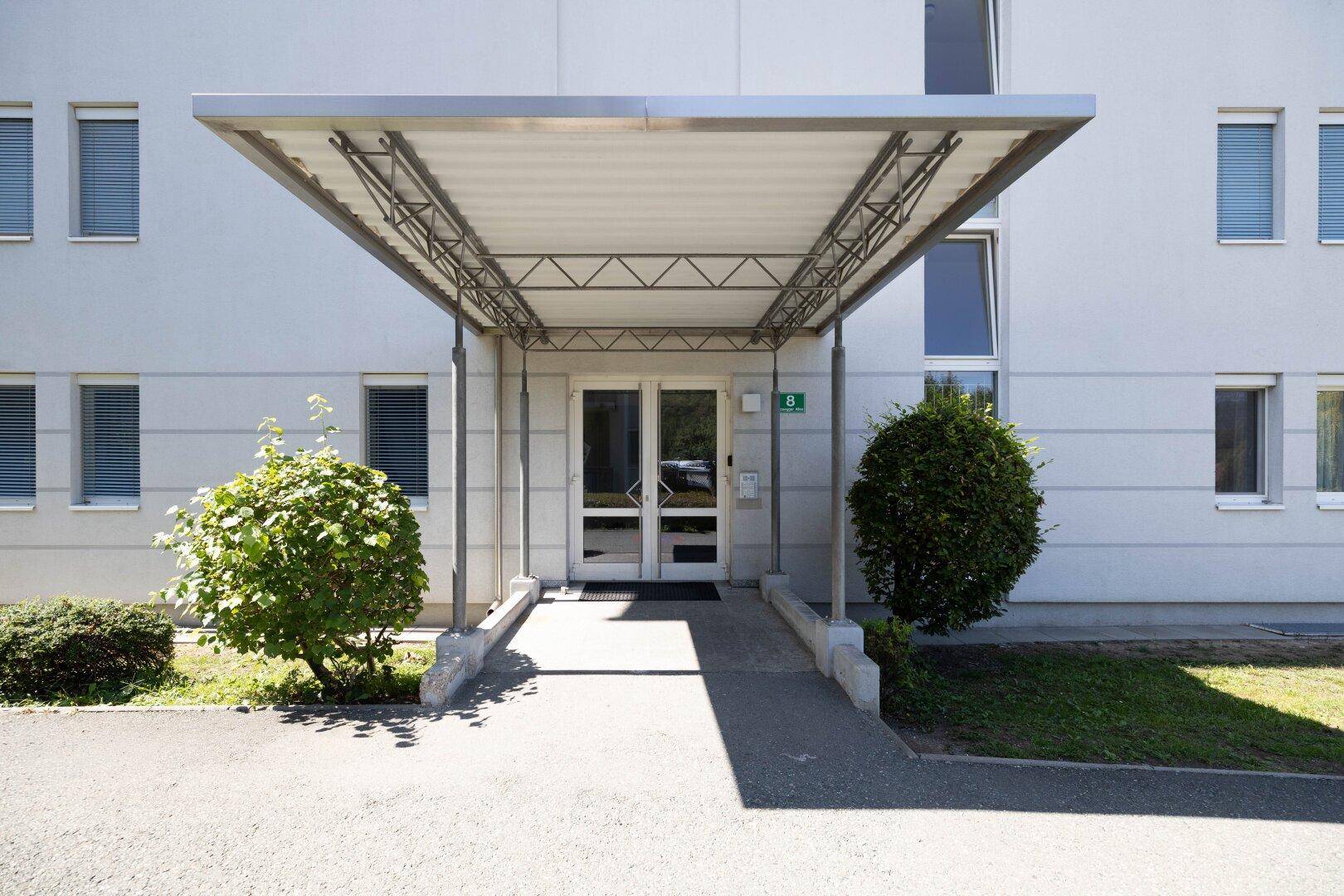 Haupteingang des Gebäudes mit schützendem Vordach und gepflegter Bepflanzung.