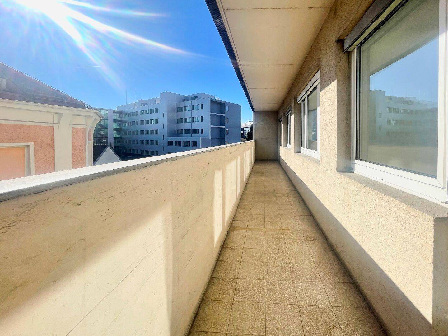 Langer sonniger Balkon mit Fliesenboden und Blick auf die umliegenden Gebäude.