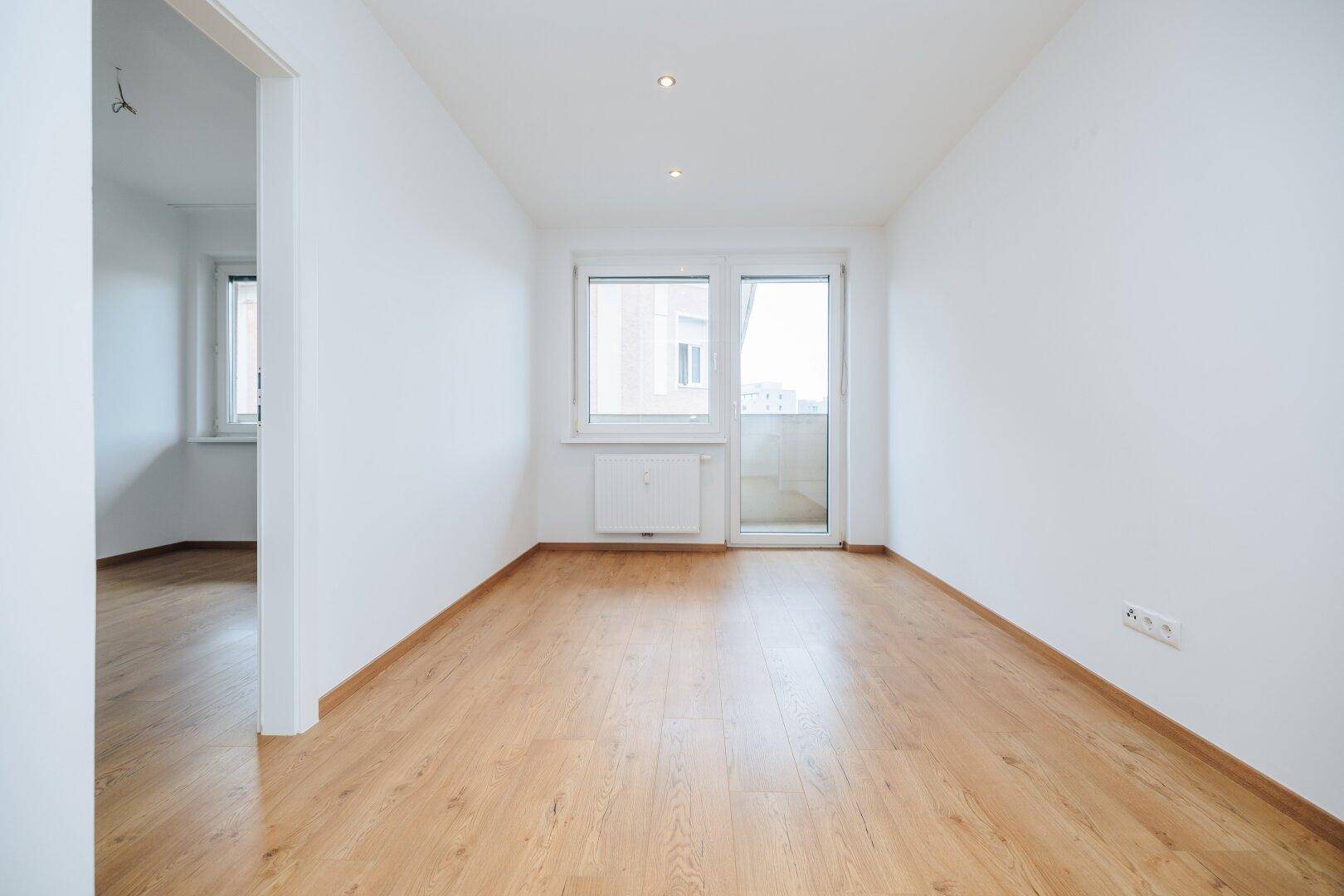 Helles leeres Zimmer mit Holzboden und Zugang zu einem Balkon oder einer Terrasse.