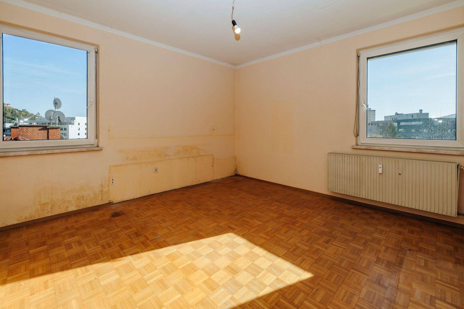 Helles Zimmer mit Parkettboden, zwei Fenstern und Blick auf die Umgebung.