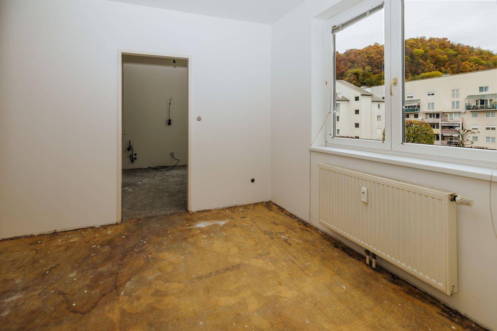 Leeres Zimmer mit Fenster und Heizkörper, angrenzend an einen weiteren Raum, mit Blick auf Bäume.