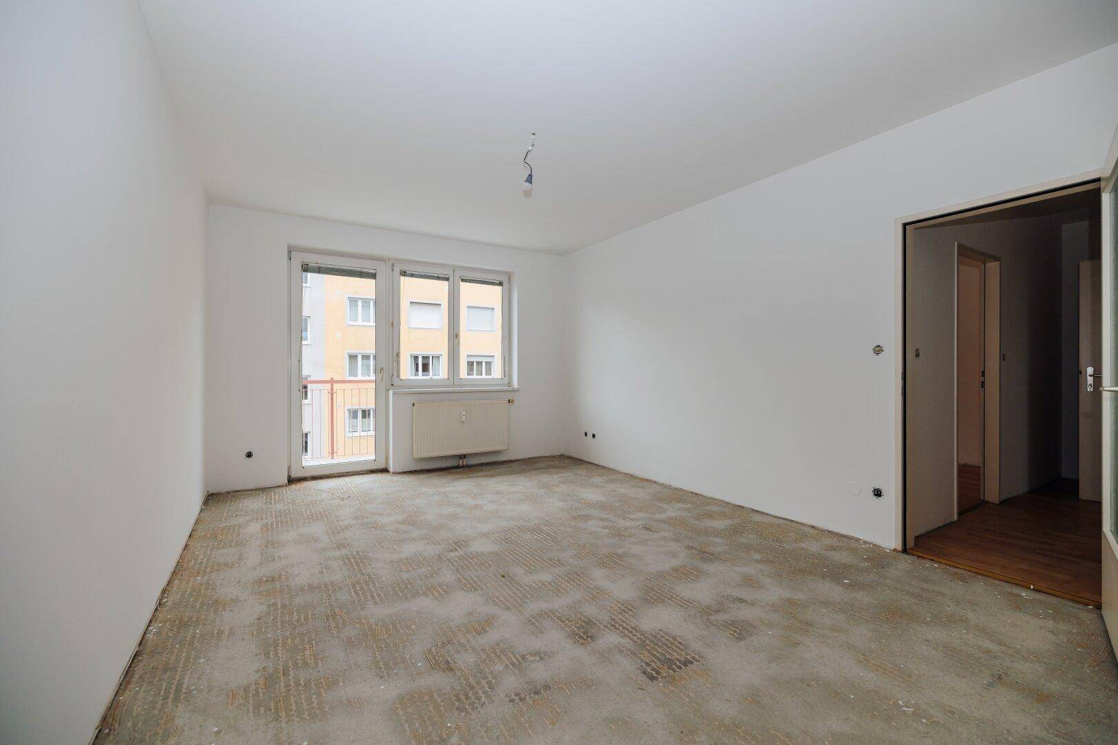 Leerer Wohnraum mit weiß gestrichenen Wänden, Fenster und Zugang zu einem Balkon, bereit für neue Böden.
