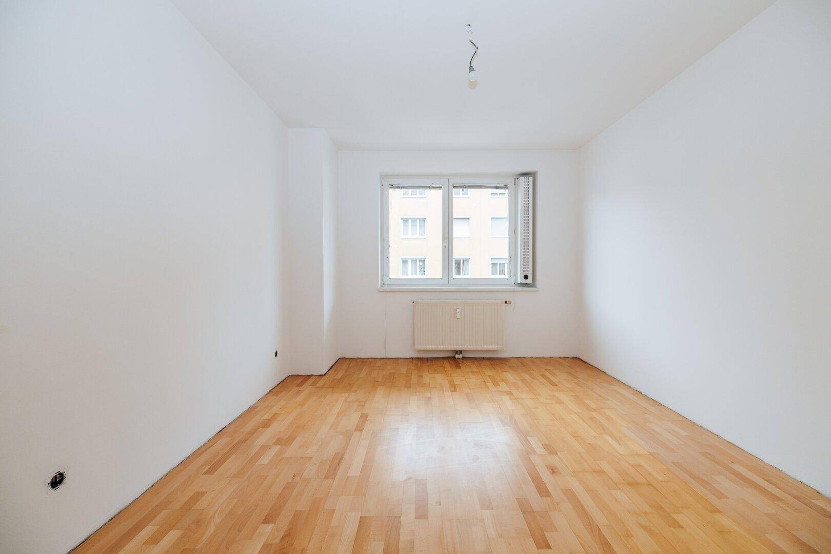 Renovierter Wohnraum mit neuem Parkettboden und großem Fenster, das viel Licht hereinlässt.
