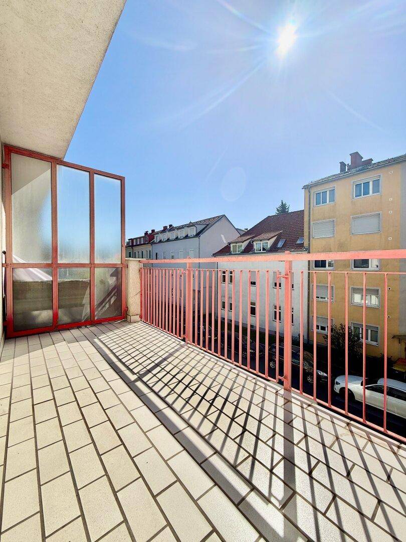 Sonniger Balkon mit rotem Geländer und Blick auf die umliegenden Gebäude und den blauen Himmel.