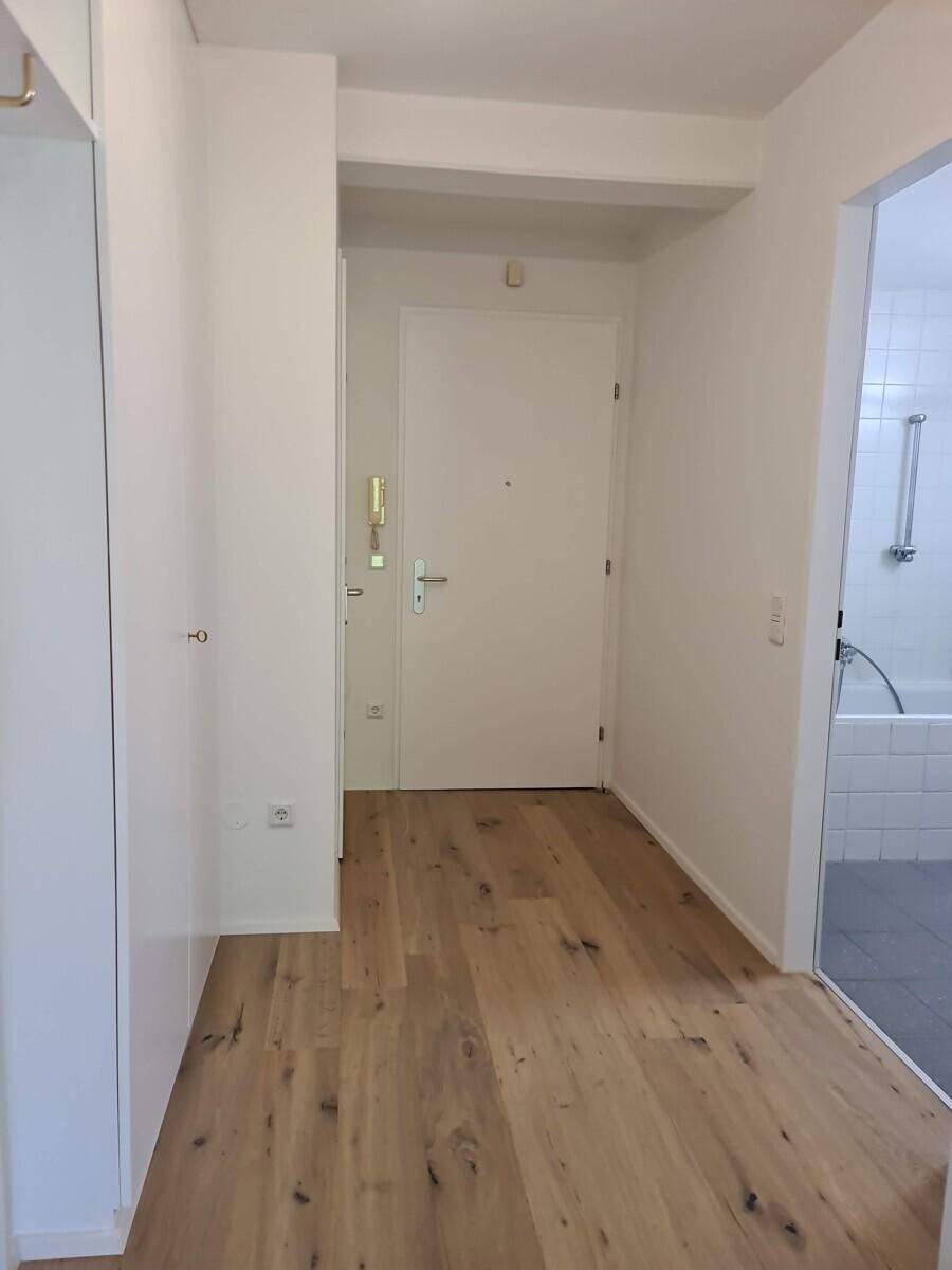 Eingangsbereich mit Holzboden, weißer Eingangstür und Blick in ein gefliestes Badezimmer.