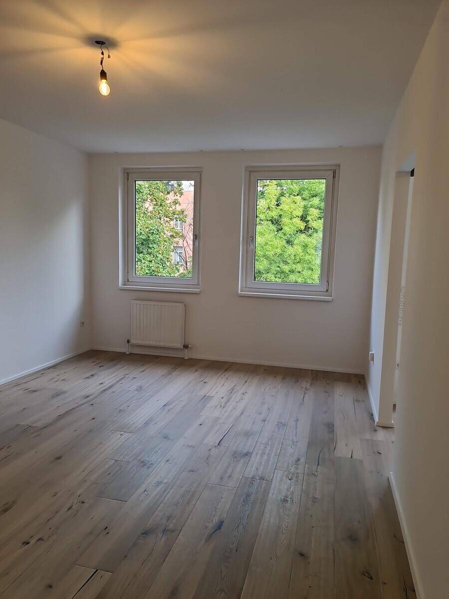 Helles Zimmer mit Holzboden und zwei Fenstern, die einen Blick ins Grüne bieten.