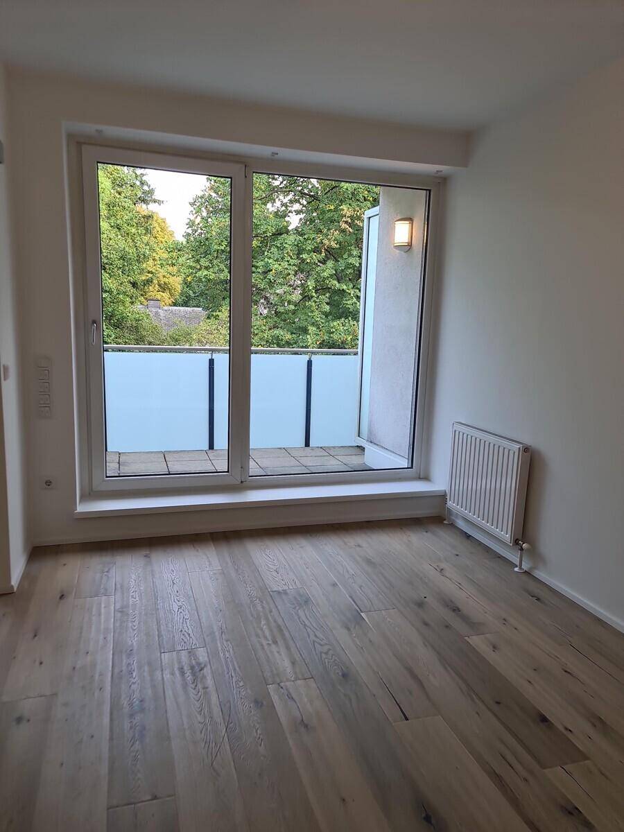Heller Raum mit Holzboden und Balkonzugang, der einen Ausblick ins Grüne ermöglicht.