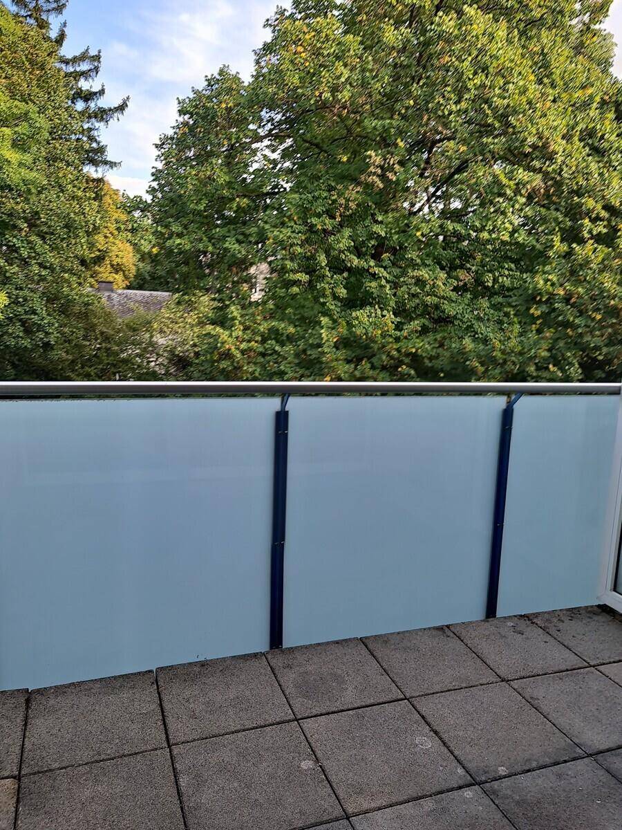 Balkon mit grauen Fliesen und Milchglasbrüstung, umgeben von grünen Bäumen.