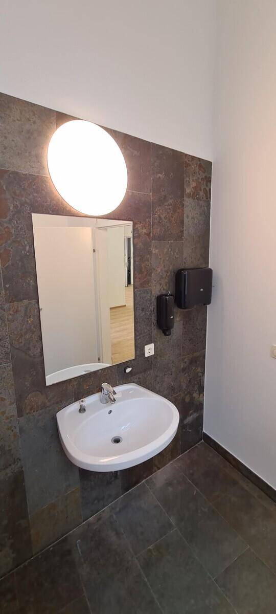 Badezimmer mit ovalem Waschbecken, beleuchtetem Spiegel und dunklen Wandfliesen.
