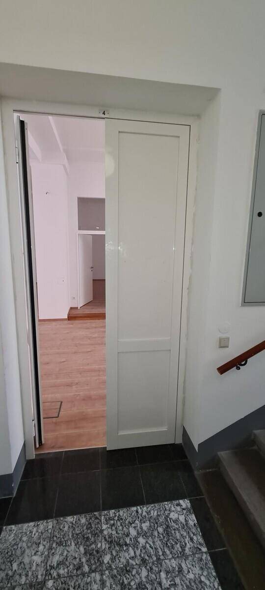 Eingangsbereich mit weißer Tür, die in einen leeren Raum führt, und Treppenhaus.