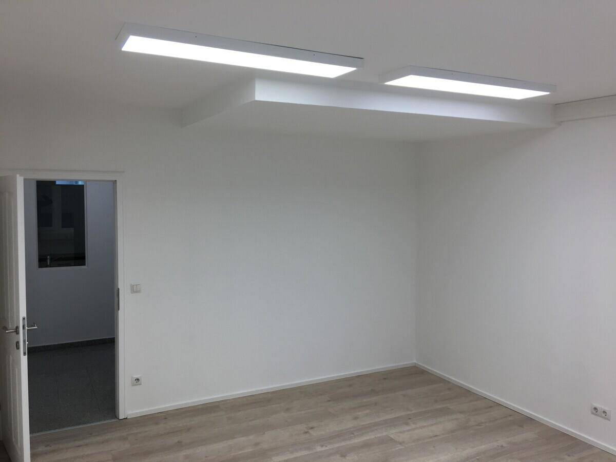 Heller Büroraum mit weiß gestrichenen Wänden, Laminatboden und offener Tür zu einem weiteren Bereich.