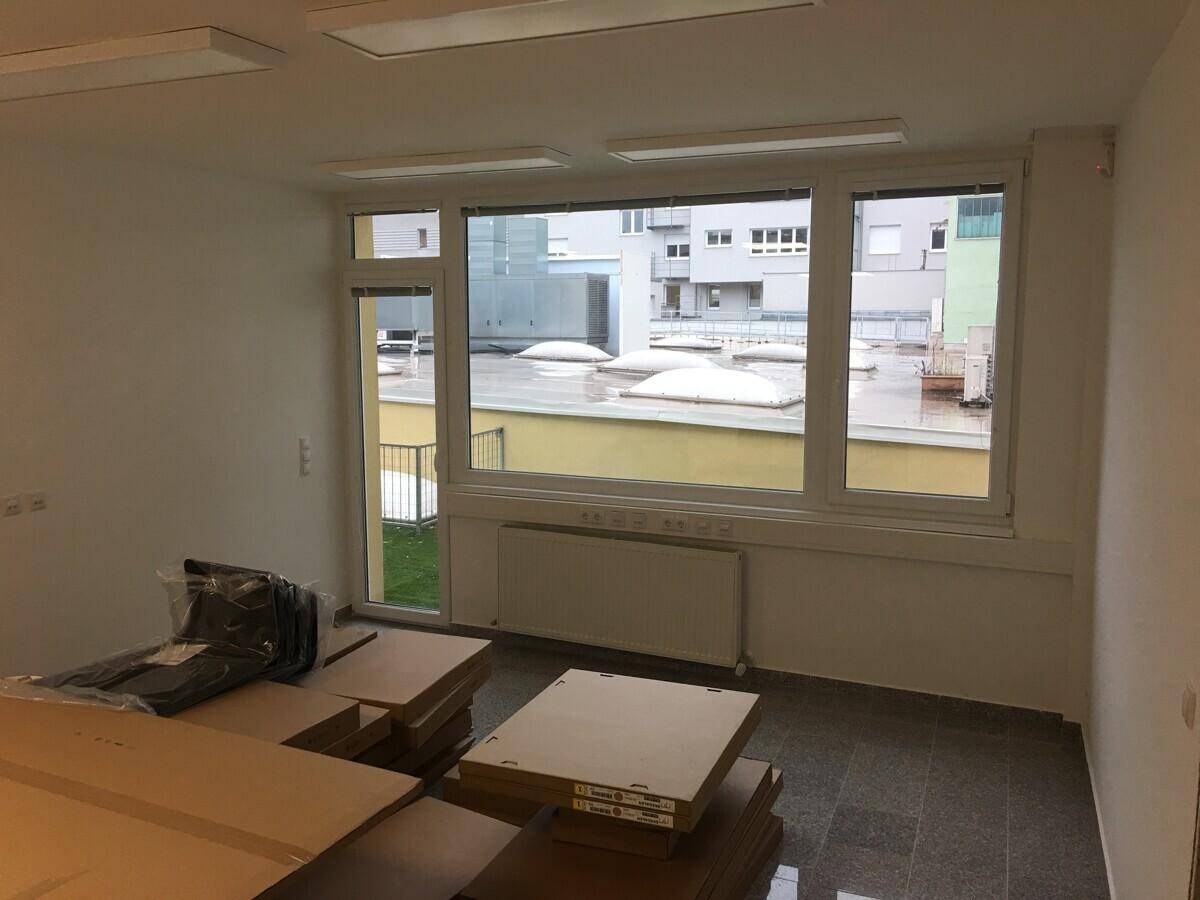 Geräumiger Büroraum mit Kartons, großen Fenstern und Blick auf eine Dachterrasse mit Oberlichtern.