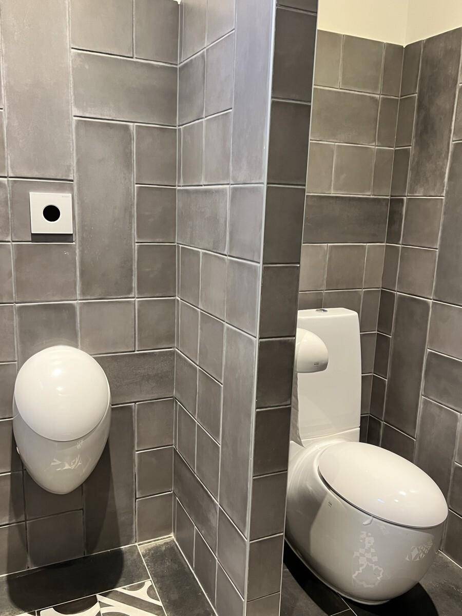 Zwei separate Toilettenkabinen mit grauen Fliesen, ausgestattet mit Urinal und Stand-WC.