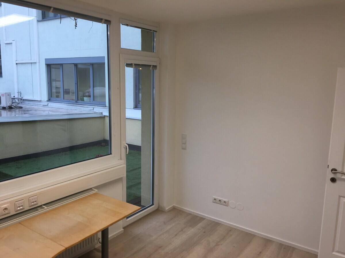 Büroraum mit Schreibtisch, großen Fenstern und Zugang zu einer kleinen begrünten Außenfläche.