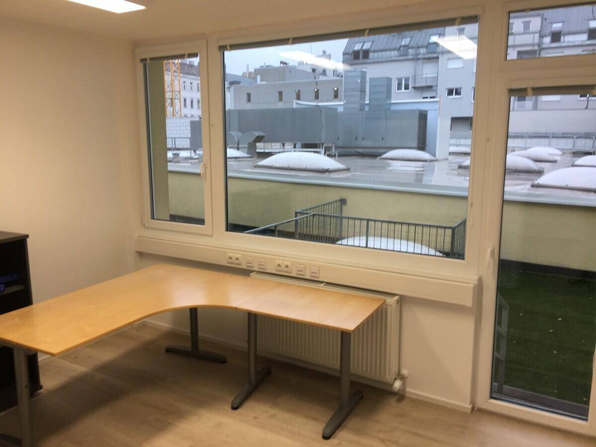Büroraum mit Schreibtisch, großen Fenstern und Blick auf eine Dachterrasse mit Kunstrasen.