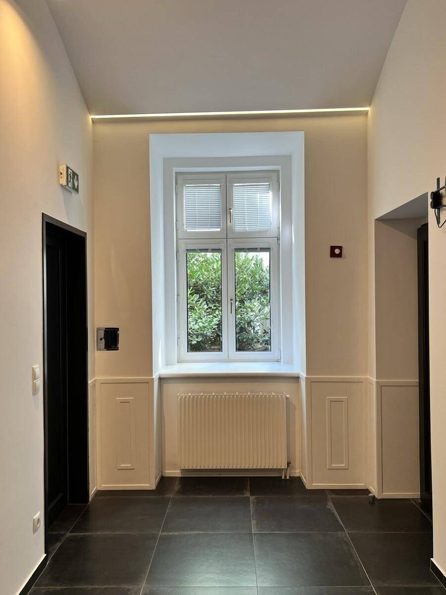 Langer Flur mit einem großen Fenster, das einen Blick ins Grüne bietet, und dunklen Türen.