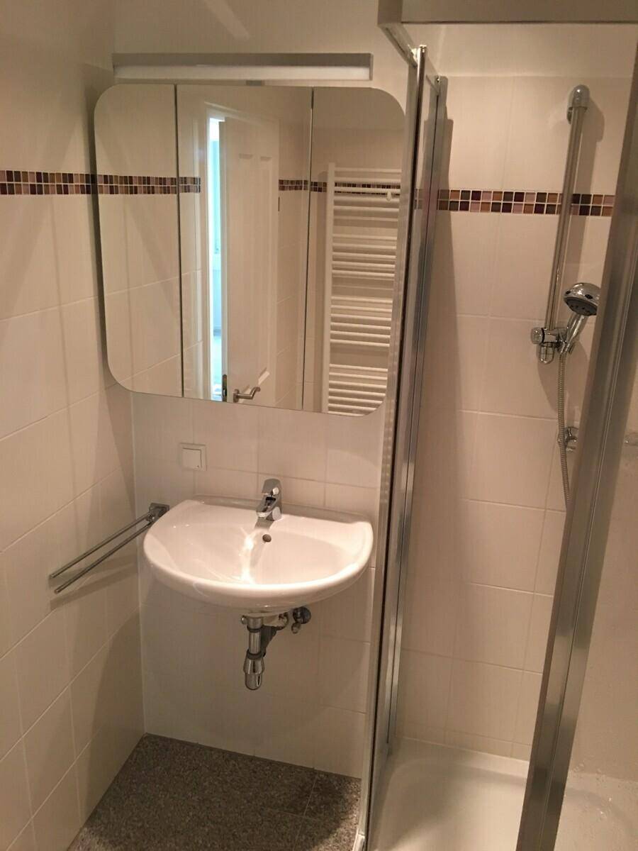 Kleines Badezimmer mit Dusche, Waschbecken, Spiegel und beigefarbenen Wandfliesen.
